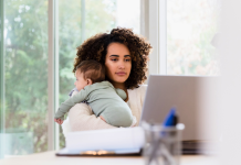 Reverse Stay-At-Home Mom working on laptop at the table with a baby in her arms.